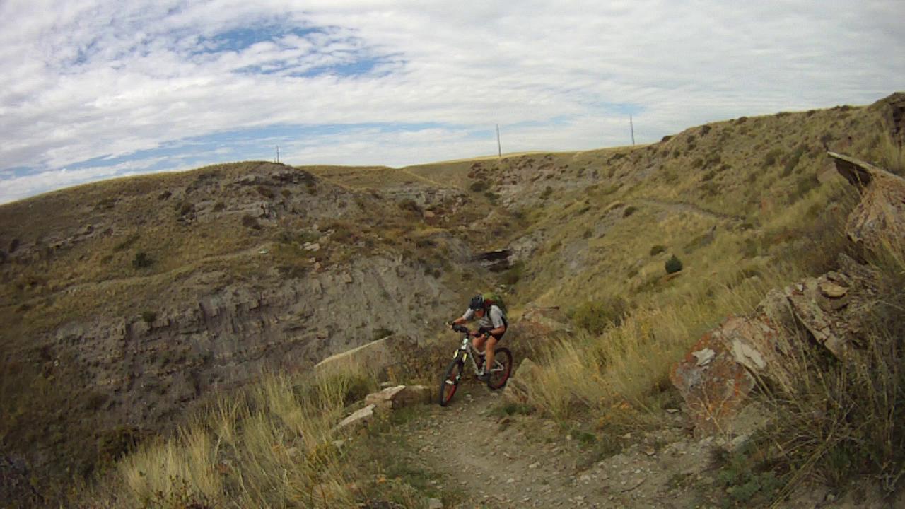 A mountain biker navigating a rocky trail in a hilly landscape under a partly cloudy sky. The terrain features tall grass and steep rocky cliffs, suggesting an adventurous outdoor setting. New Video Game/Yeah Kid mountain bike trail.