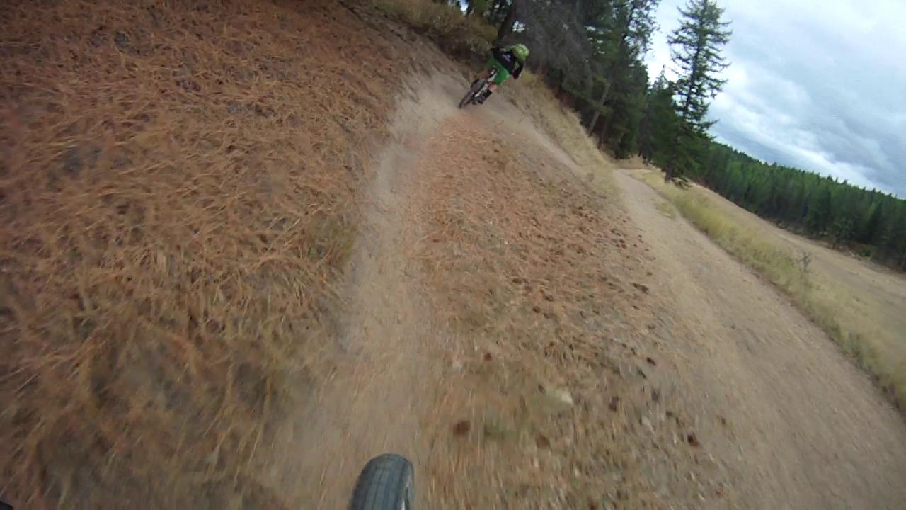 A mountain biker navigates a winding dirt trail surrounded by pine trees, with a focus on the bike's front wheel and the path ahead. The terrain is covered in fallen pine needles, and the sky overhead is partly cloudy, suggesting an adventurous ride in a natural setting. Rattlesnake mountain bike trail.
