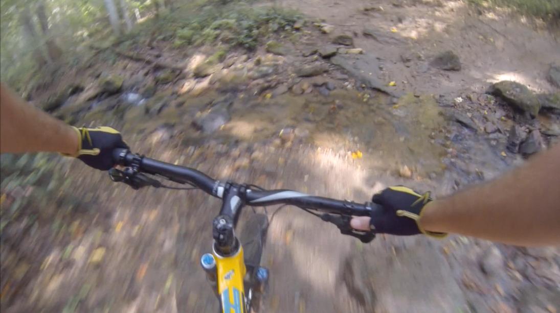 Alt text: A view from above the handlebars of a mountain bike navigating a rocky trail with patches of dirt and small water streams, surrounded by greenery. Gunpowder Falls State Park mountain bike trail.