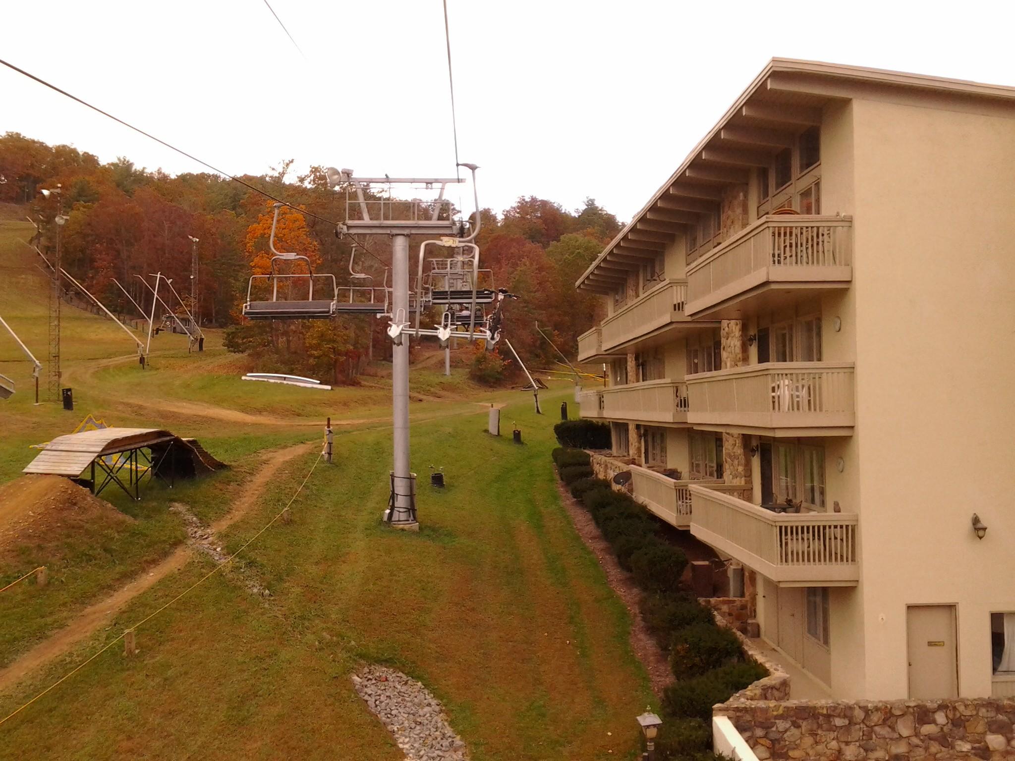 Mongoose Blackcomb: A ski resort scene featuring a chairlift in the foreground, surrounded by green grass and autumn trees with colorful foliage in the background. To the right, a multi-story hotel with balconies overlooks the ski area, with a gravel path and grassy slopes leading towards the lift.
