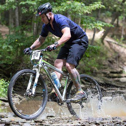 Airborne Goblin: A cyclist in a helmet rides a mountain bike through a stream, splashing water as he navigates rocky terrain in a lush forest setting. He is wearing a blue shirt and black shorts, with visible tattoos on his arms.