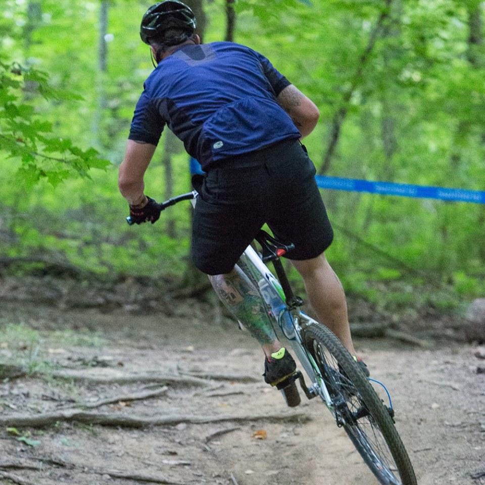 Airborne Goblin: A cyclist riding a mountain bike on a forest trail, surrounded by green trees and foliage. The rider is seen from behind, wearing a blue jersey and black shorts, as they navigate a curving path. The image highlights the natural setting and the action of mountain biking.