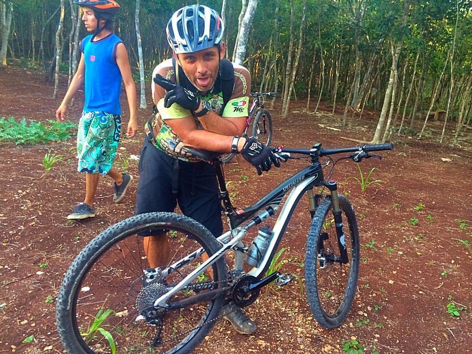 Specialized Camber Expert Carbon 29: Two individuals are seen outdoors in a forested area. One person, wearing a helmet and gloves, is posing with a mountain bike and making a playful expression. The other person, dressed in a blue tank top and patterned shorts, stands in the background, looking at the camera. The ground is brown and earthy, with some greenery in the surroundings.