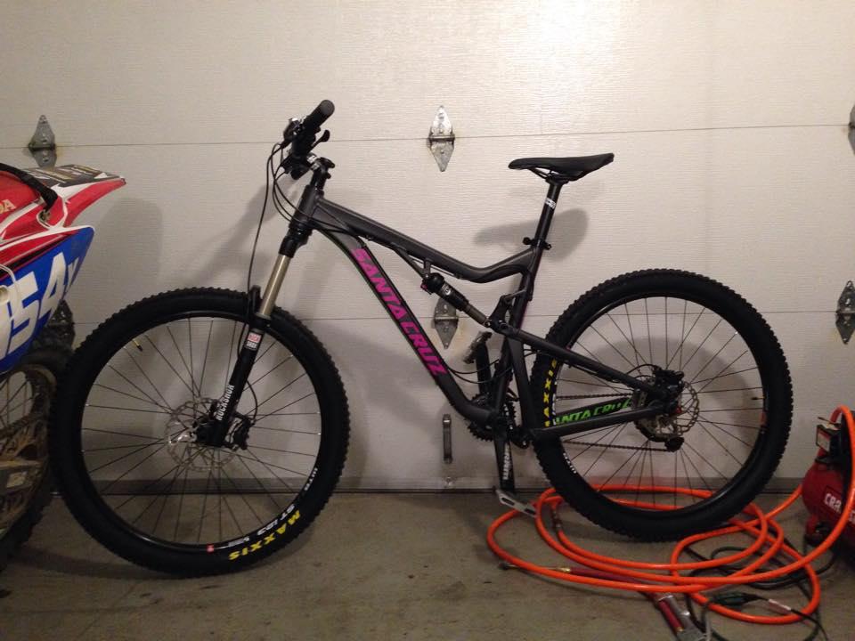 Santa Cruz Bronson: A black Santa Cruz mountain bike with pink and green accents, parked next to a red motocross bike in a garage setting. The garage door is visible in the background, and there are coiled hoses on the floor.