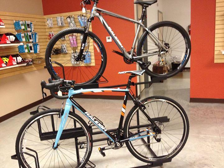 Two bicycles displayed in a bike shop; one is a silver mountain bike on the top rack, and the other is a black and blue road bike on the bottom rack. The background features shelves with various cycling accessories and apparel.