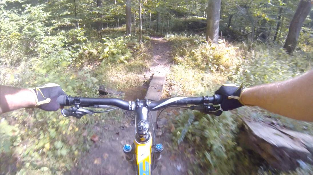 First-person view of a mountain biking trail, showing handlebars and a rider's arms wearing gloves as they navigate a wooded path. The scene features lush greenery and a narrow wooden bridge over a small gap on the trail. Gunpowder Falls State Park mountain bike trail.
