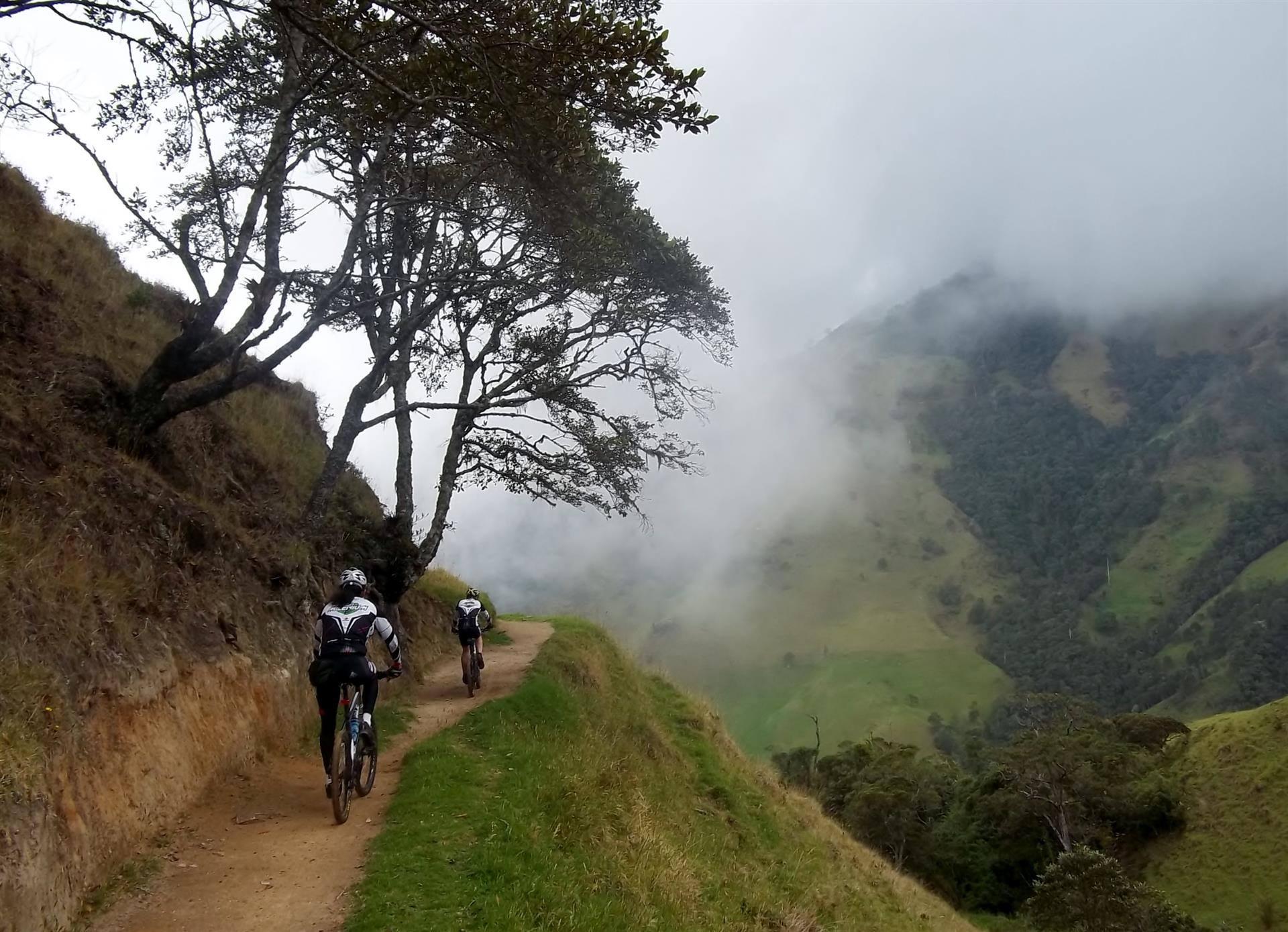 Two mountain bikers riding along a narrow dirt path on a misty hillside, surrounded by lush green vegetation and towering trees. The landscape features rolling hills partially obscured by fog, creating a serene and adventurous setting. Peñas Blancas mountain bike trail.