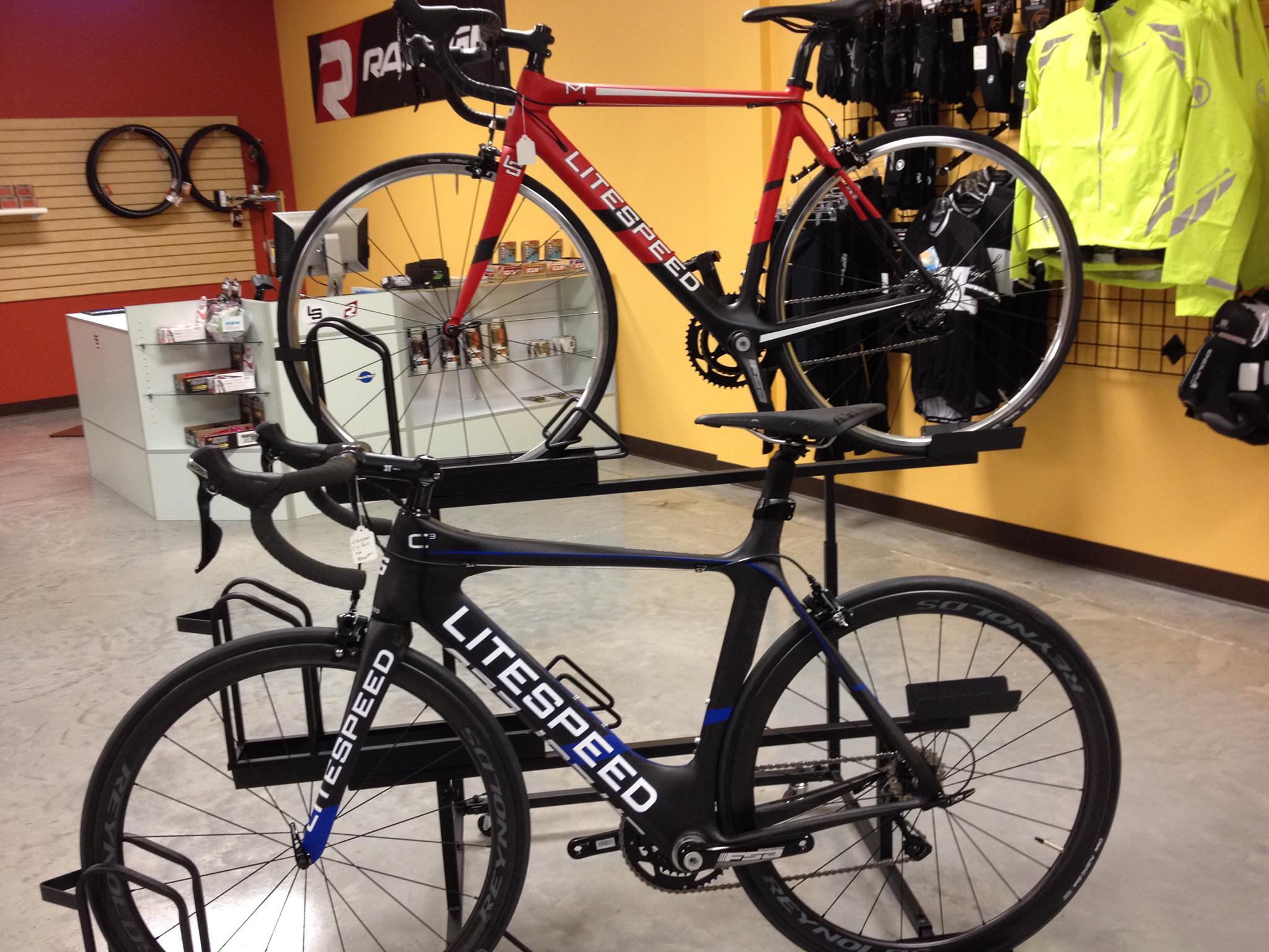 Image of two Litespeed bicycles displayed in a bike shop. The upper bike is red and the lower bike is black, both featuring sleek designs and racing-style handlebars. The background includes shelves stocked with bike accessories and clothing.