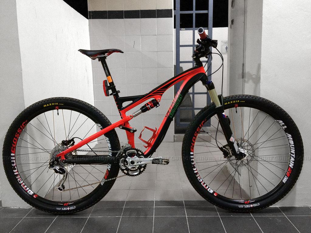 Specialized Camber 29: A side view of a red and black mountain bike positioned indoors against a tiled wall. The bike features thick tires labeled "Maxxis," a sleek frame with suspension, and a comfortable seat. The background includes a door and a grid pattern on the wall.