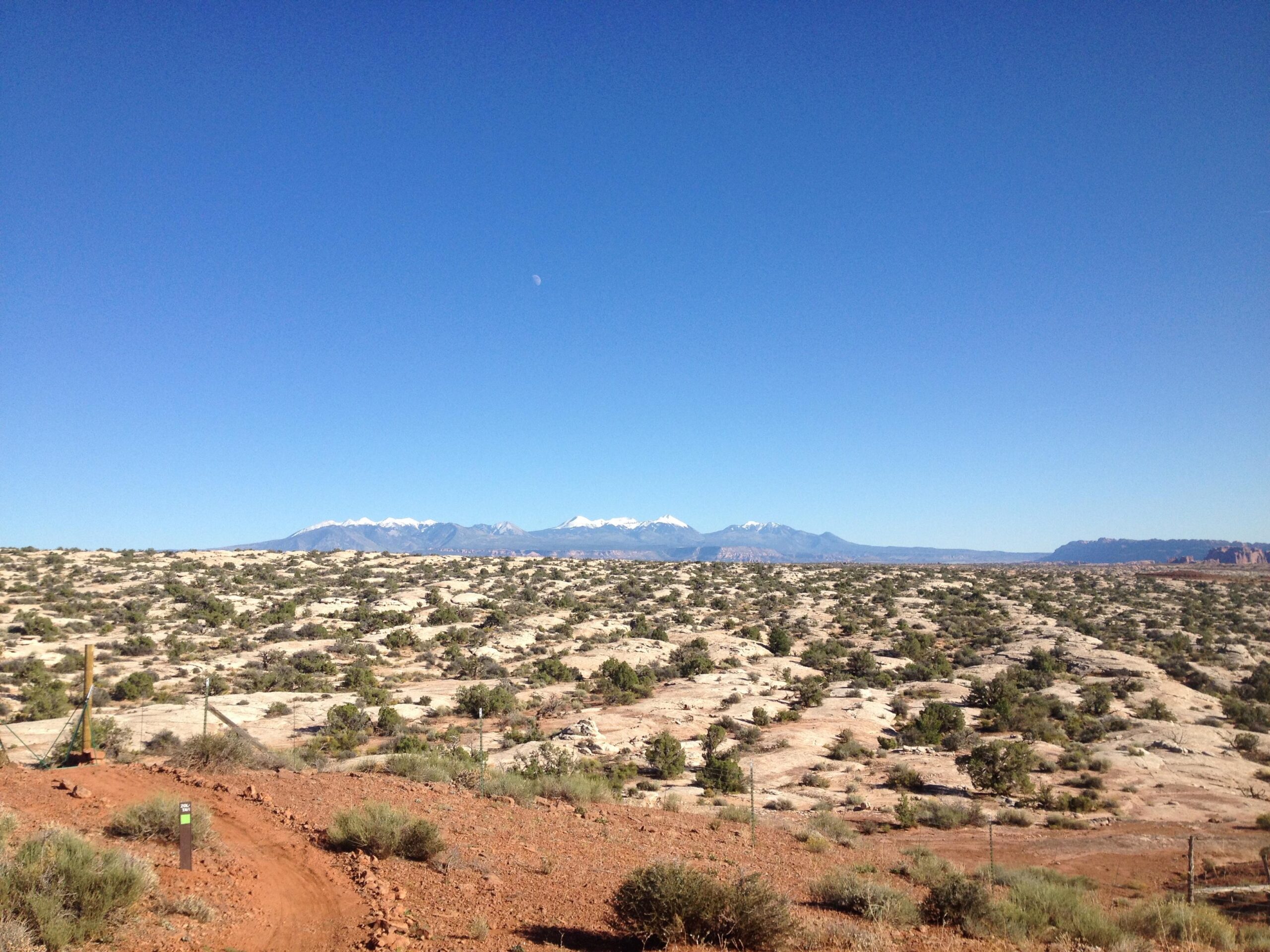 A panoramic view of a desert landscape with scattered shrubs and rocky terrain, leading to distant snow-capped mountains under a clear blue sky. A winding dirt path and a signpost are visible in the foreground, while the moon can be seen in the sky. Zoltar mountain bike trail.