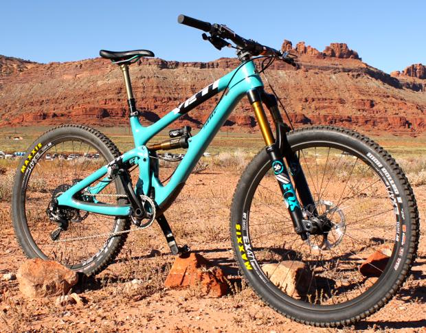 Yeti SB6: A turquoise mountain bike positioned on rocky terrain with red desert cliffs in the background under a clear blue sky.