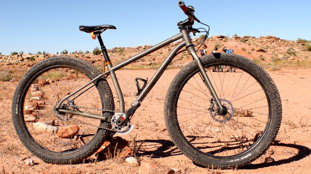 Niner ROS 9 Plus: A mountain bike with fat tires is positioned on rocky terrain under a clear blue sky. The bike has a metallic frame and features a single-speed setup, making it suitable for off-road rides. In the background, there are more rocky hills and sparse vegetation, suggesting a rugged outdoor environment.