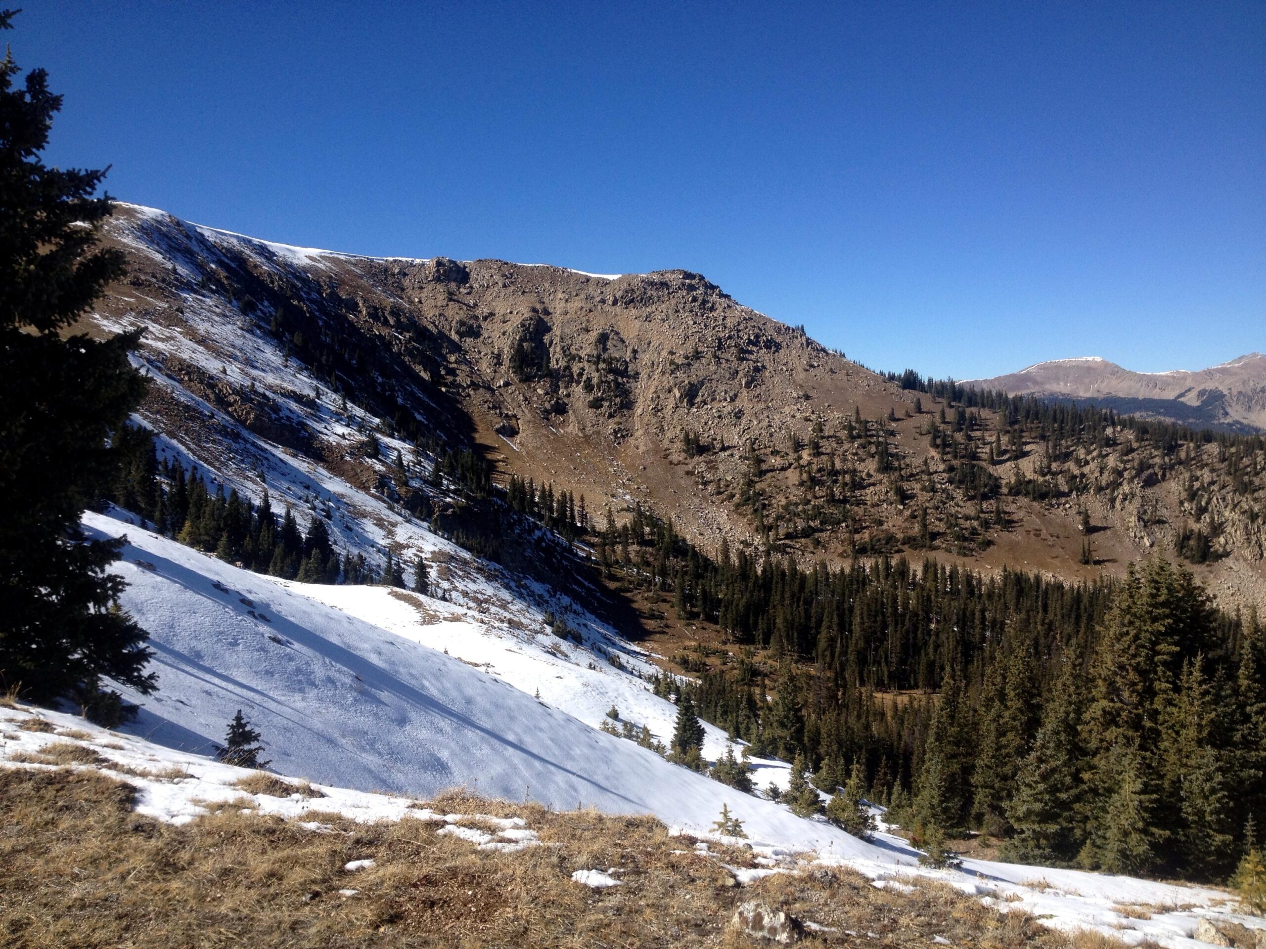 A scenic mountainous landscape featuring a mix of rocky terrain and patches of snow, with evergreen trees dotting the lower slopes. In the background, steep hills rise under a clear blue sky, showcasing the rugged beauty of the natural environment. Monarch Crest Trail mountain bike trail.