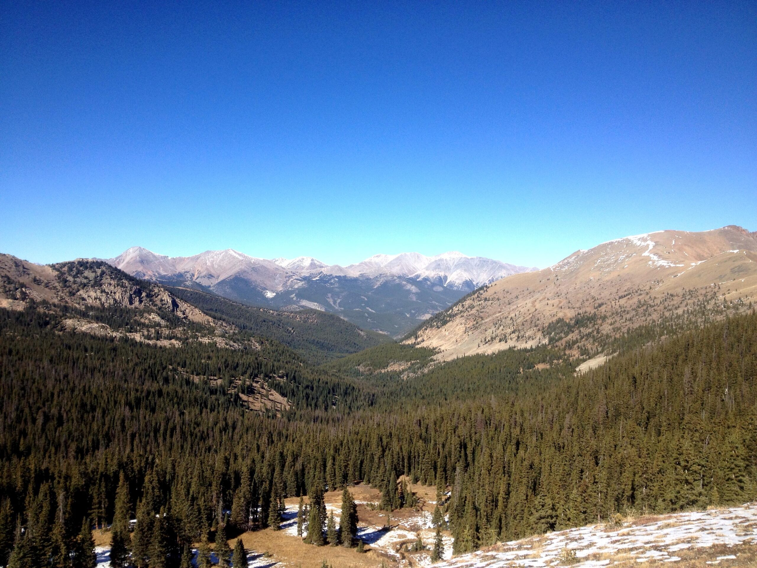 A panoramic view of a mountainous landscape under a clear blue sky, showcasing rugged peaks in the background and dense evergreen forests in the foreground. Snow can be seen on some mountaintops, and patches of snow are visible on the ground, suggesting a late winter or early spring setting. Monarch Crest Trail mountain bike trail.