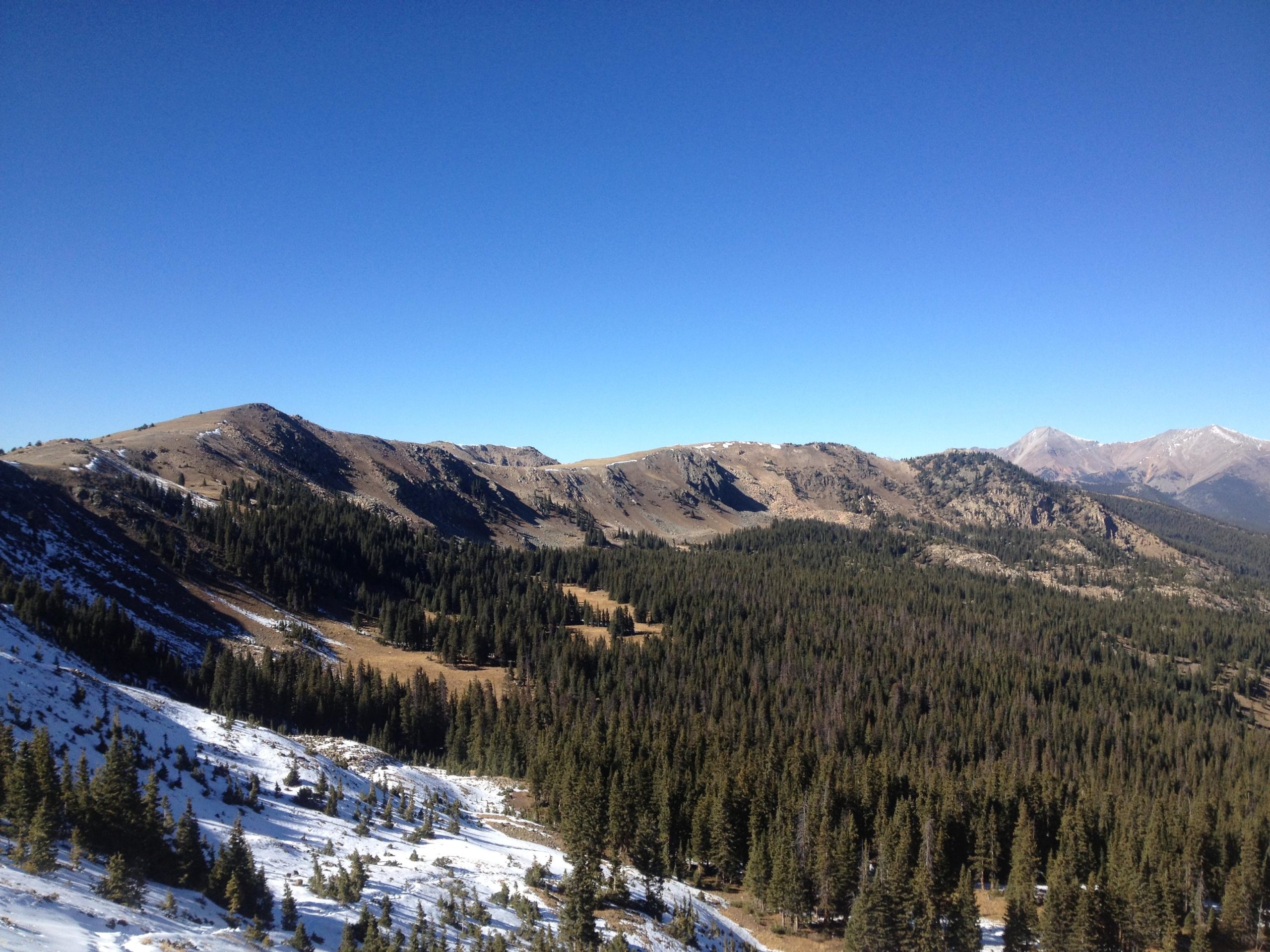 A panoramic view of mountainous terrain featuring rolling hills, coniferous forests, and patches of snow, under a clear blue sky. Monarch Crest Trail mountain bike trail.