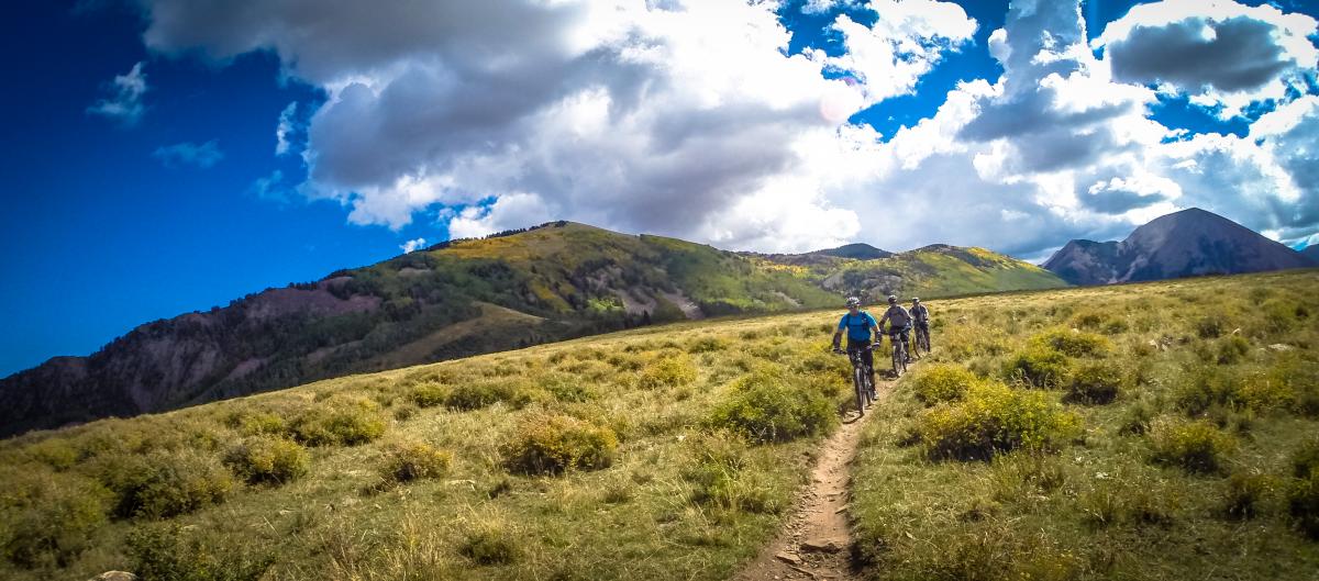 Three mountain bikers riding along a dirt trail in a grassy mountain landscape under a blue sky filled with clouds. Rolling hills and peaks are visible in the background, showcasing a vibrant mix of green and autumn colors.