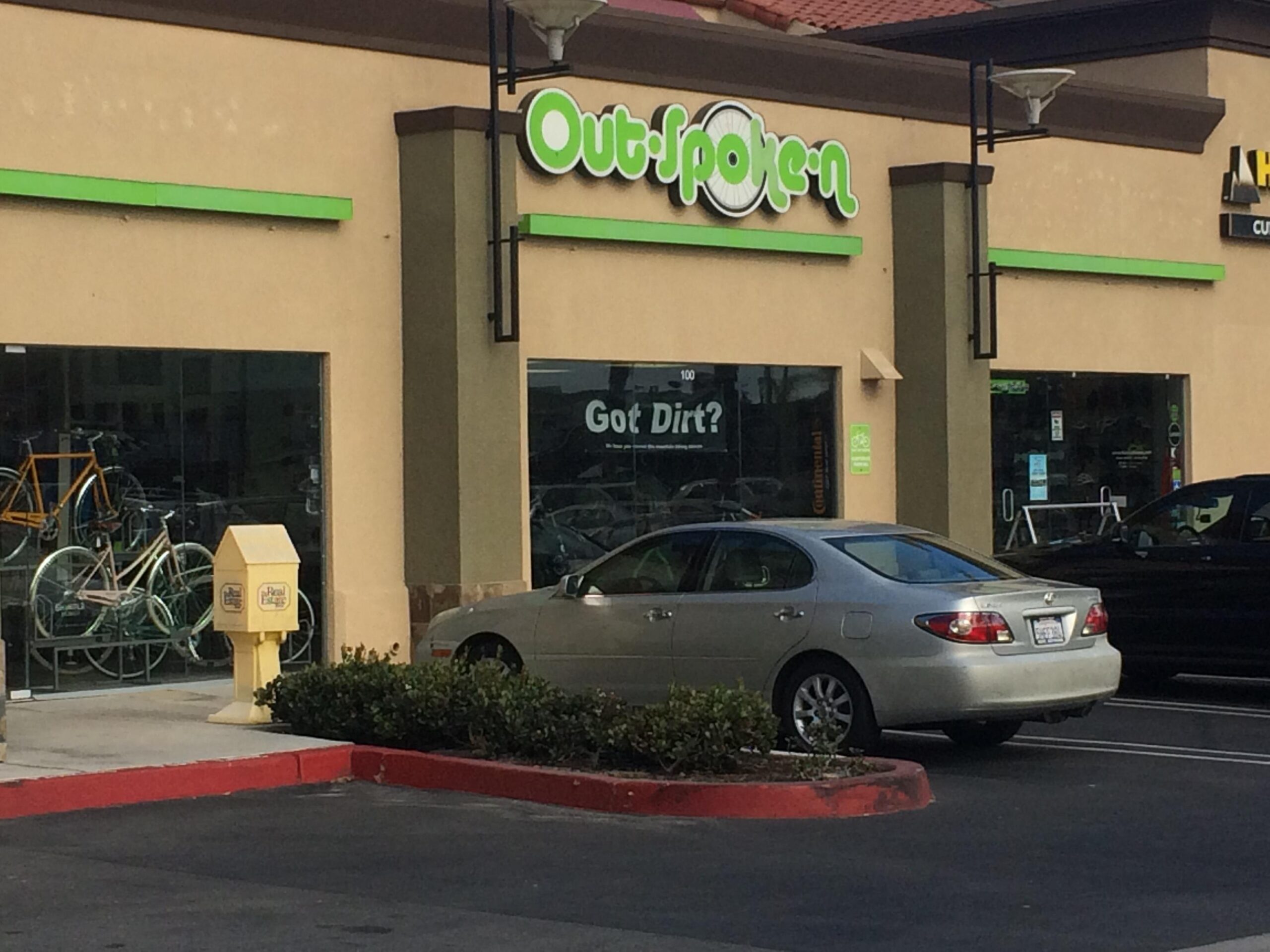 Image of the storefront of "Outspoken," featuring a large sign above the entrance. The window displays bicycles, and a sign reads "Got Dirt?" in the window. A yellow newspaper box is positioned near the entrance, and a silver car is parked in front of the store. The building has a beige exterior with green accents.