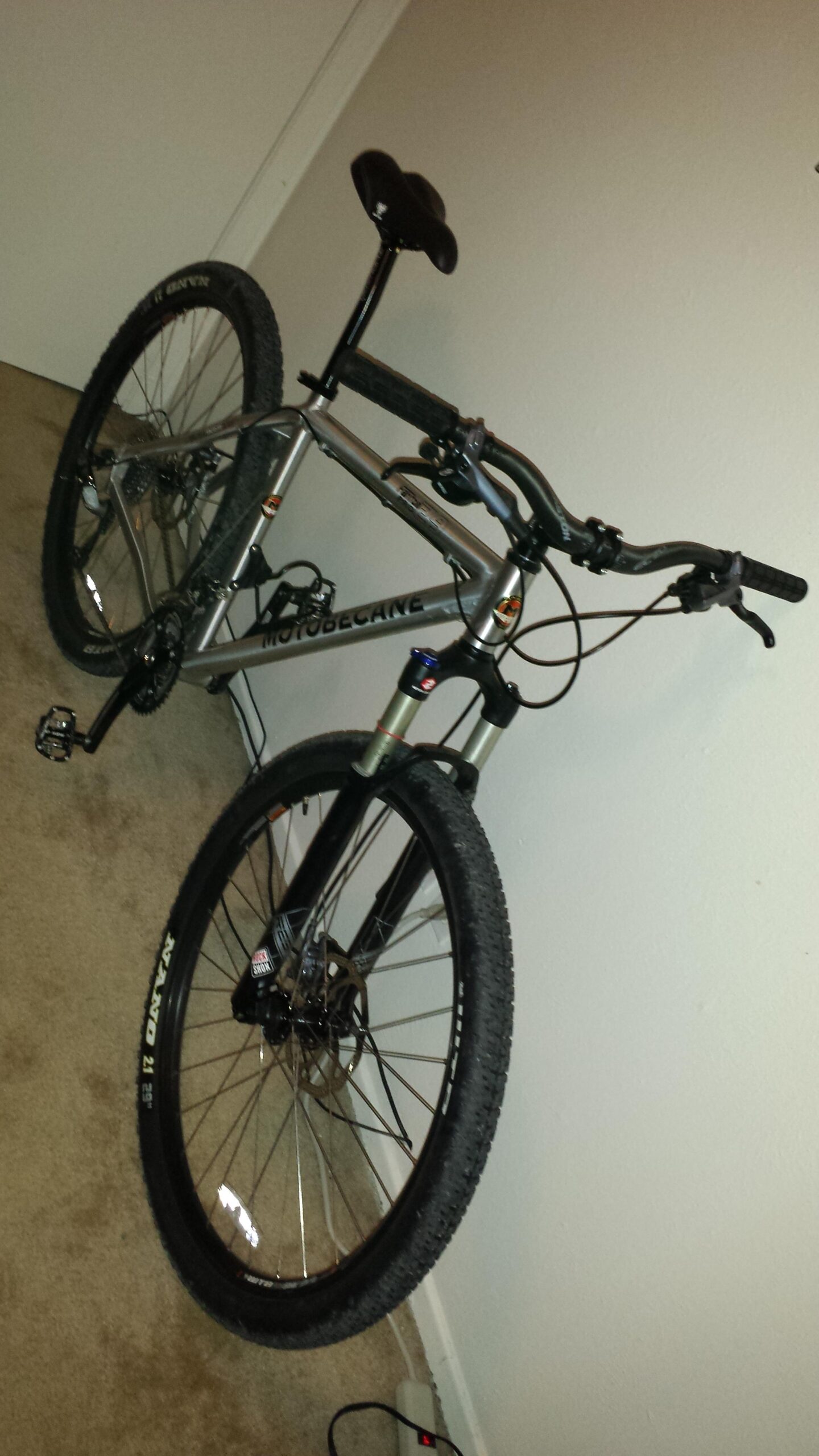 Motobecane Fantom 29PRO: Alt text: A Motobecane mountain bike stands upright against a wall, featuring a silver frame, black handlebars, and thick tires. The bike is positioned on a carpeted floor, with a power strip visible in the foreground.