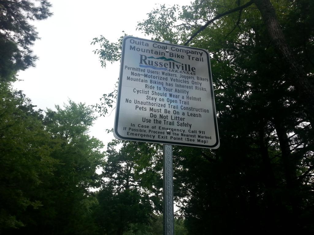 Sign for the Ouitta Coal Company Mountain Bike Trail in Russellville, Arkansas. The sign lists permitted users, safety recommendations, and rules for trail use, including requirements for helmets and leash laws for pets. Surrounding trees and greenery are visible in the background. Sweet Spot Trail (Ouita Coal Company Trail) mountain bike trail.