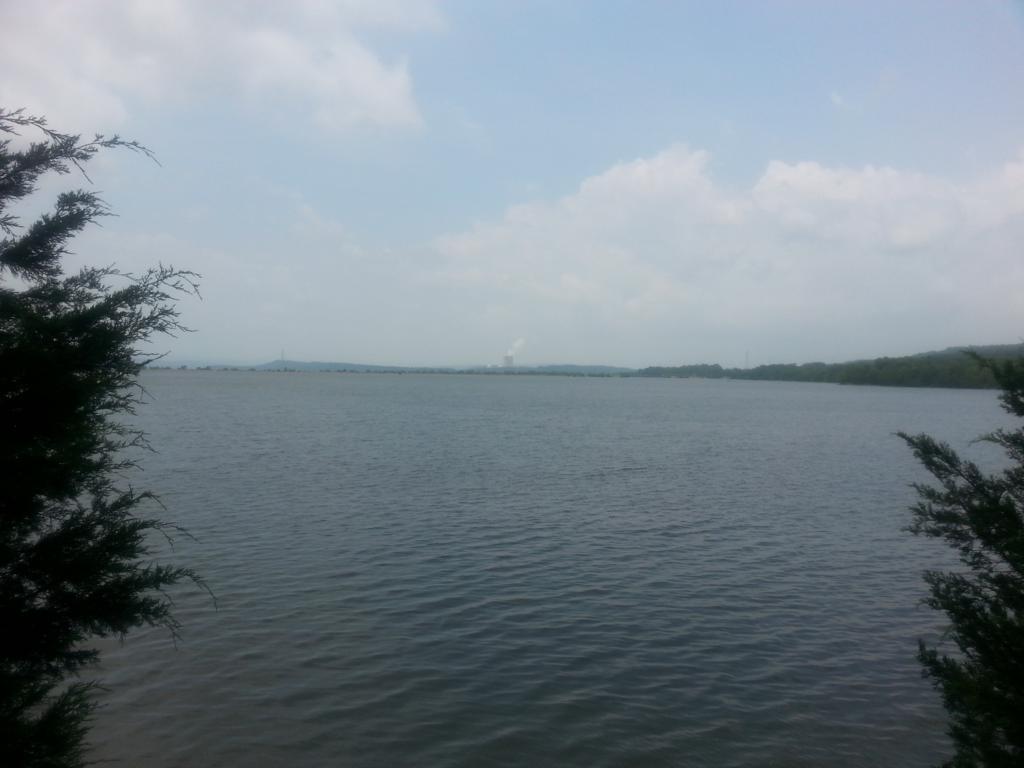 A tranquil view of a lake surrounded by trees, under a partially cloudy sky. In the distance, hills and a smokestack are visible, indicating human activity nearby. Sweet Spot Trail (Ouita Coal Company Trail) mountain bike trail.