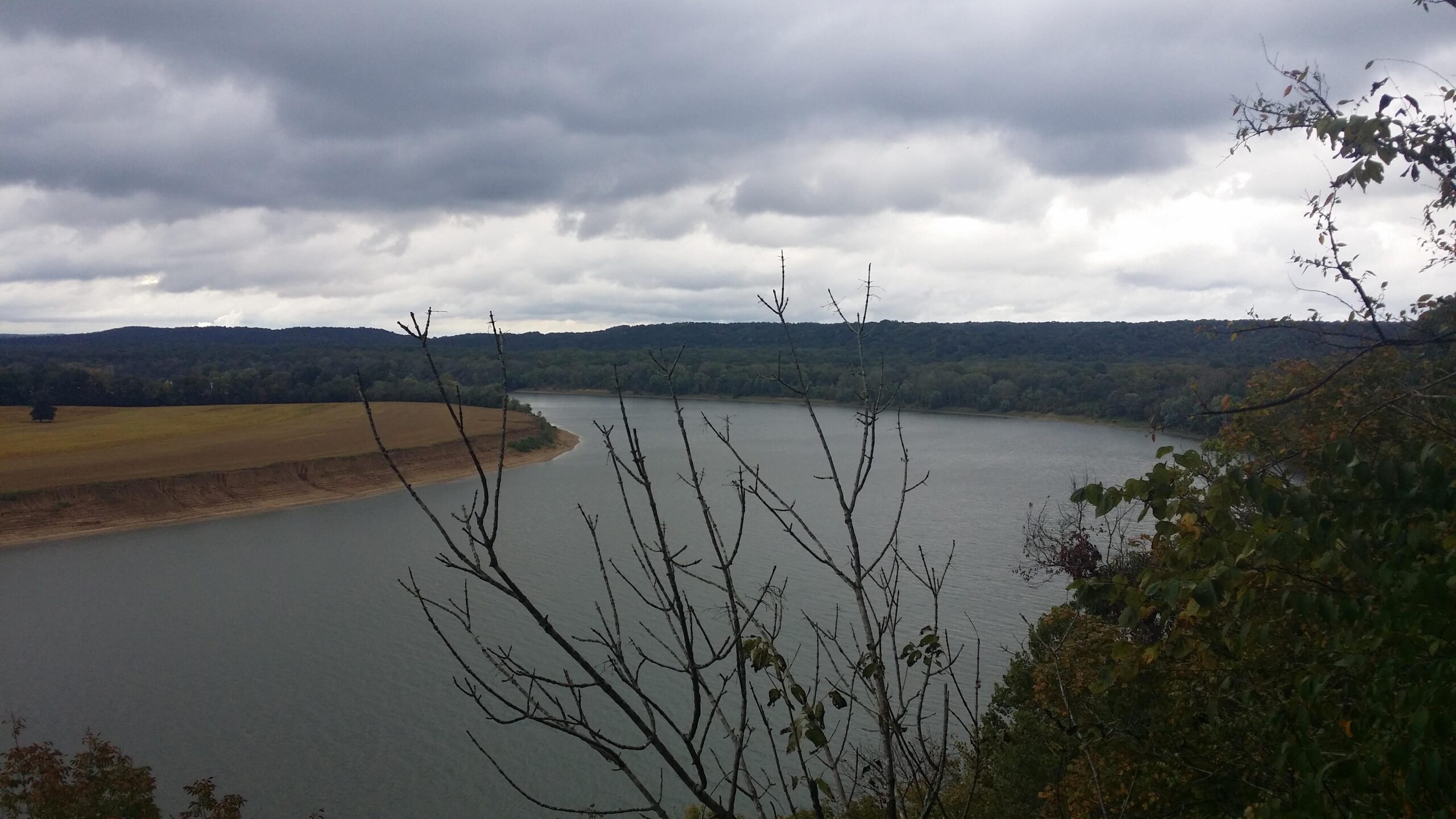 A serene view of a river winding through a landscape under a cloudy sky, with barren trees in the foreground and rolling hills in the background. The water has a calm surface reflecting the overcast atmosphere. Otter Creek mountain bike trail.