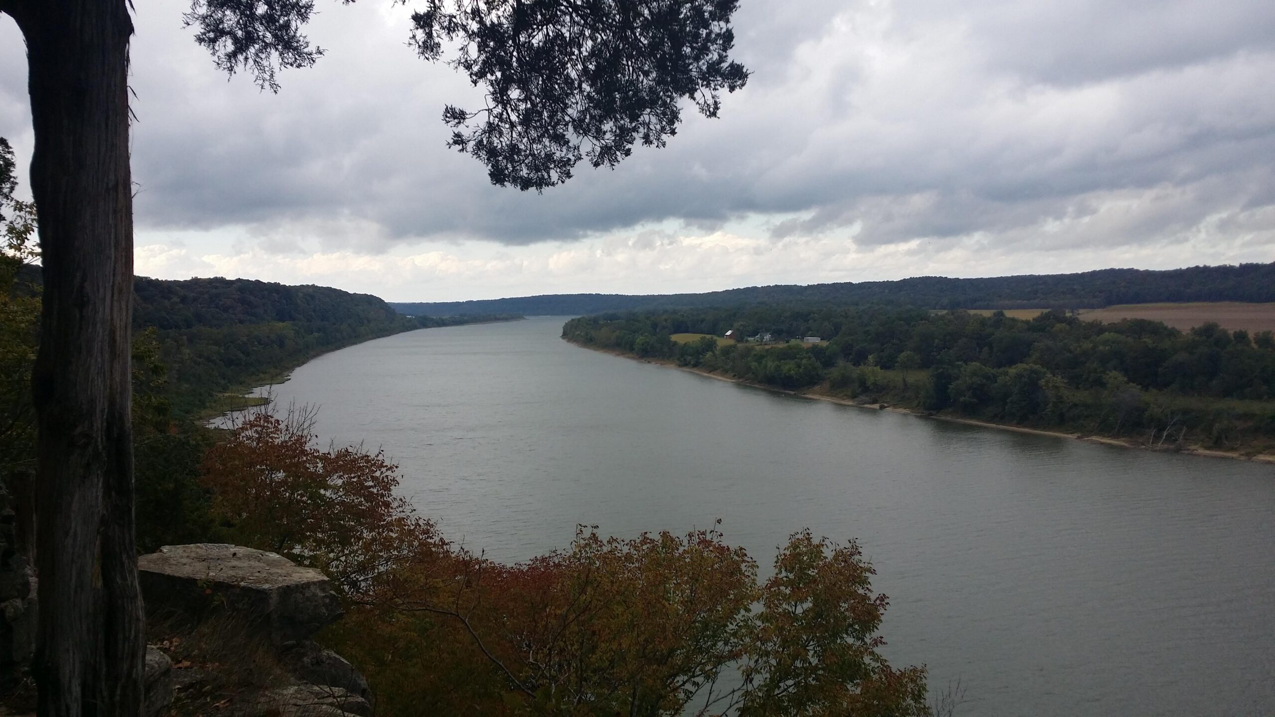 A scenic view of a river meandering through lush green landscapes under a cloudy sky. The foreground features trees with autumn foliage, and the riverbank displays gentle slopes leading to fields and distant hills in the background. Otter Creek mountain bike trail.