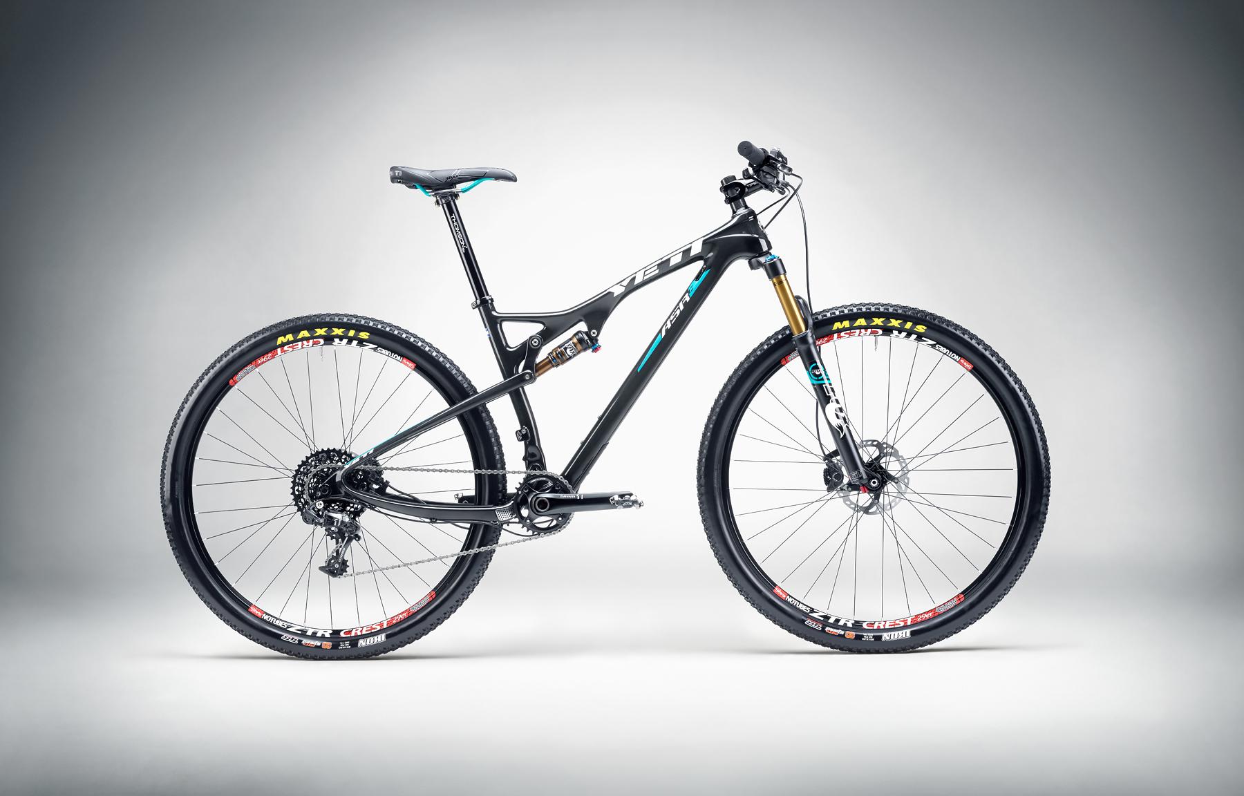 Yeti ASRc: A black mountain bike with blue accents, featuring a lightweight frame and front suspension, positioned on a gradient background. The bike is equipped with Maxxis tires and a 1x drivetrain setup.