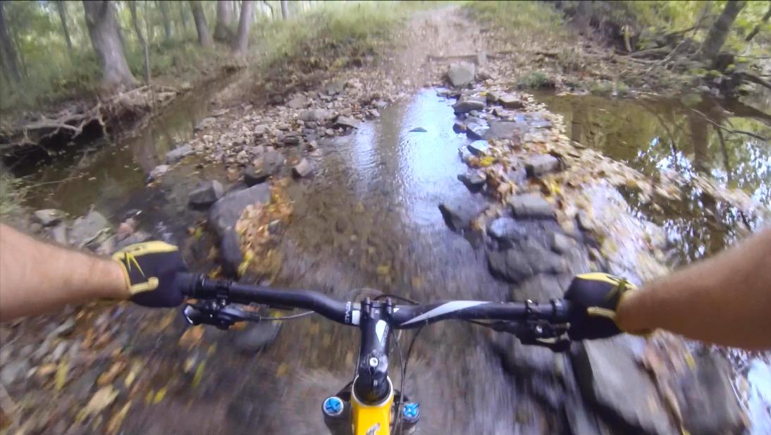 A cyclist's hands gripping the handlebars of a mountain bike, navigating a rocky path with a shallow stream running across it, surrounded by trees and fallen leaves. Gunpowder Falls State Park mountain bike trail.