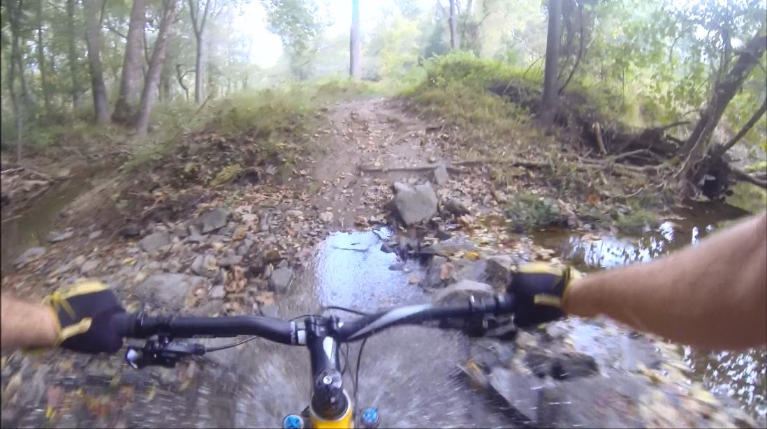 A mountain biker's perspective riding through a rocky trail with a small stream running along the path, surrounded by trees and foliage in a natural setting. Gunpowder Falls State Park mountain bike trail.