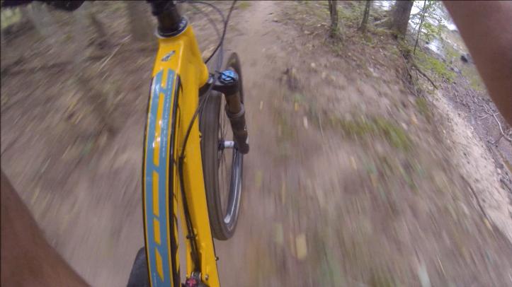 A close-up view of a bicycle tire on a dirt trail, with trees and natural surroundings blurred in the background, suggesting motion. The bike features a bright yellow frame and a partially visible handlebar. Gunpowder Falls State Park mountain bike trail.