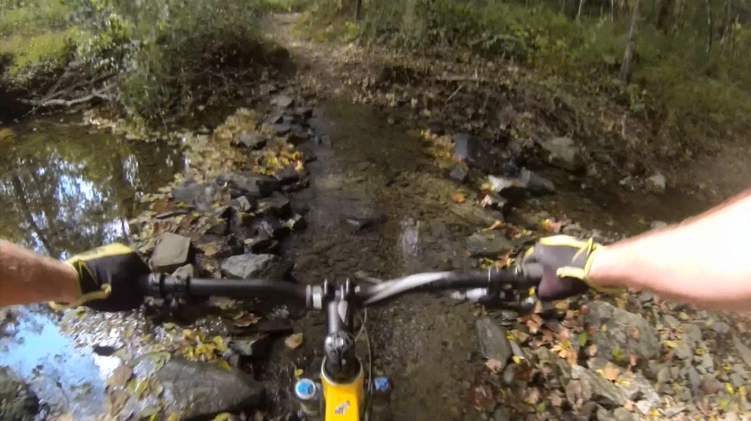 A view from the handlebars of a mountain bike navigating a rocky path beside a small stream, with autumn leaves scattered on the ground and trees lining the trail. Gunpowder Falls State Park mountain bike trail.