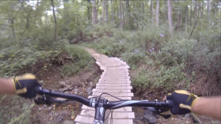 A view from a mountain bike handlebar, showing a wooden bridge over a small stream in a forested area. The path is surrounded by green foliage and trees, suggesting an outdoor biking trail. The rider's hands are visible, wearing gloves, gripping the handlebars as they navigate the narrow bridge. Gunpowder Falls State Park mountain bike trail.