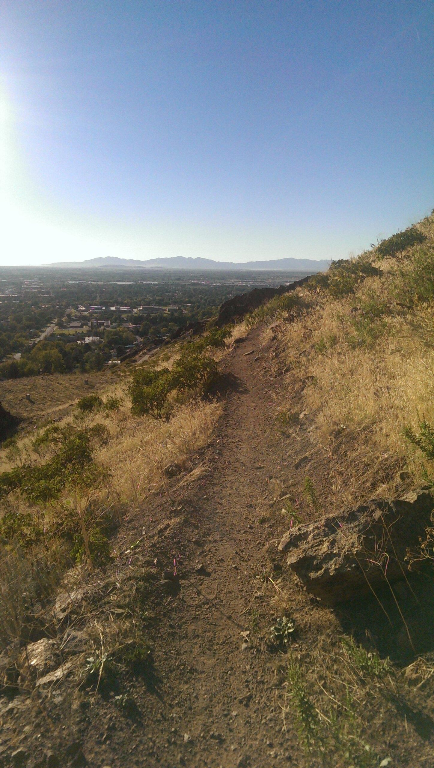 A narrow dirt hiking trail winds along a hillside, surrounded by dry grass and small shrubs. In the distance, a view of a valley and mountains under a clear blue sky is visible. The sunlight is shining brightly, creating a serene outdoor atmosphere. Bonneville Shoreline Ogden North Of 12th mountain bike trail.