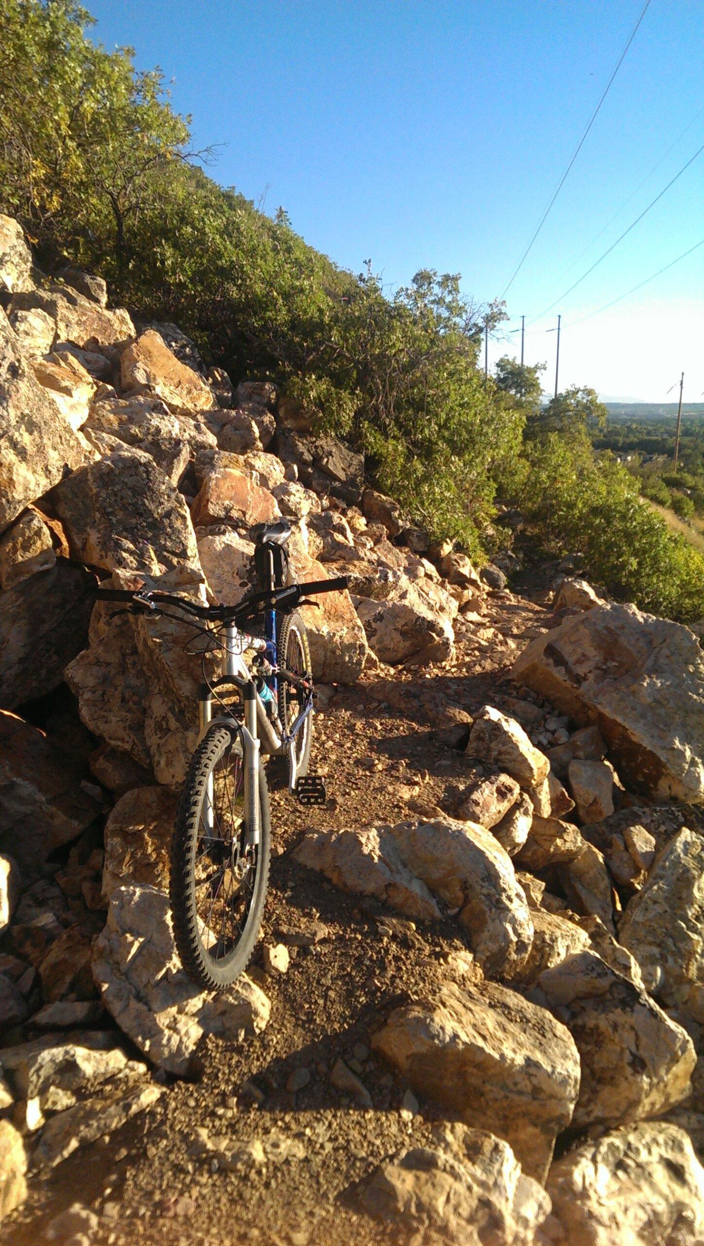 A mountain bike positioned on a rocky trail surrounded by greenery and a clear blue sky. The bike leans against a large rock amidst smaller pebbles, suggesting an adventurous outdoor setting. Bonneville Shoreline Ogden North Of 12th mountain bike trail.