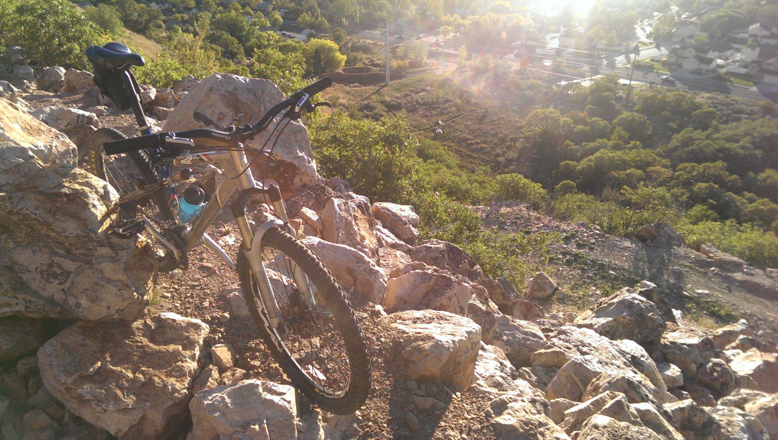A mountain bike resting on rocky terrain with a view of a lush valley and distant houses under a sunny sky. The sunlight creates a warm glow over the landscape, highlighting the details of the rocks and foliage. Bonneville Shoreline Ogden North Of 12th mountain bike trail.