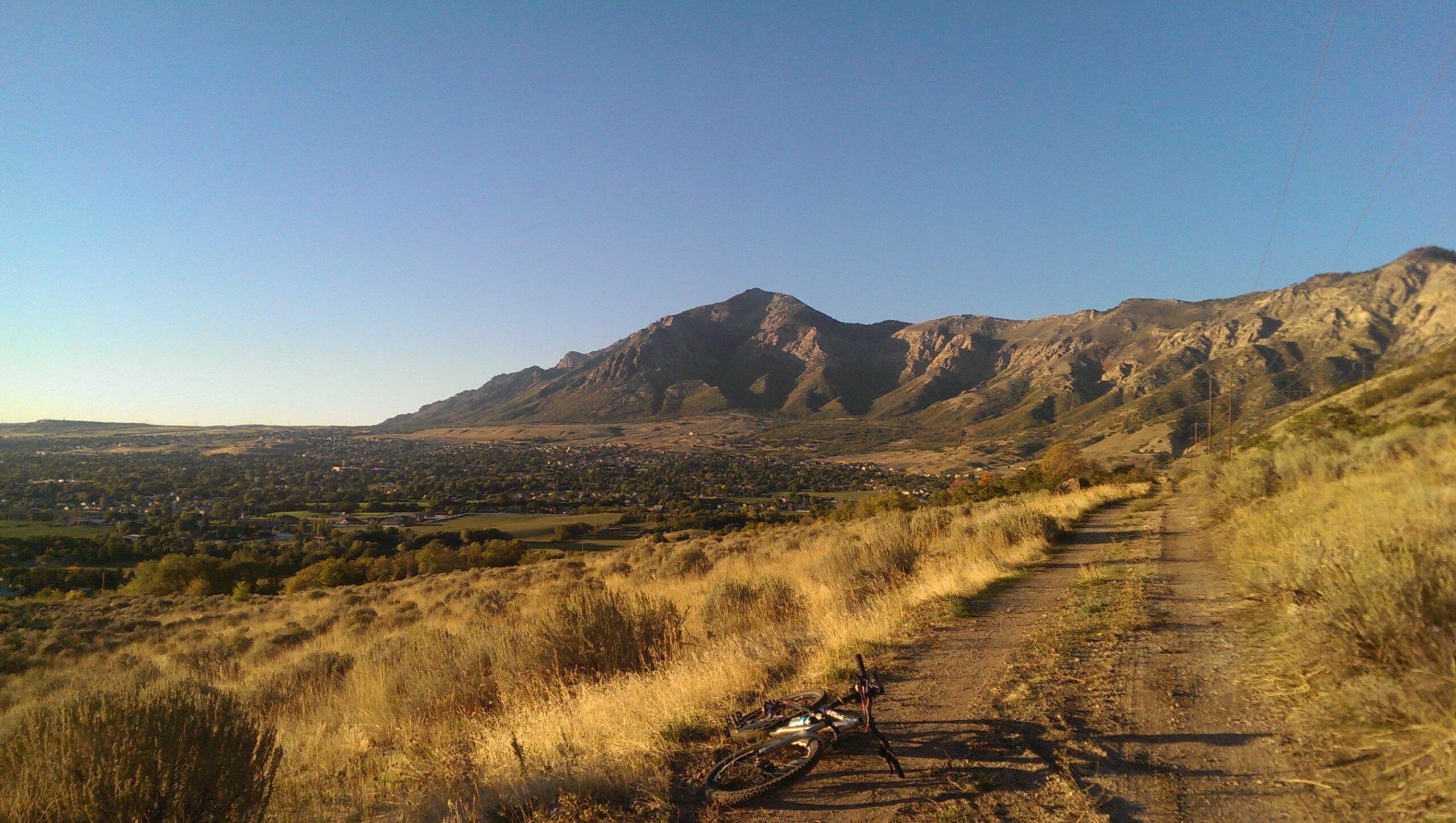 A scenic view of a mountainous landscape with gentle slopes, framed by a clear blue sky. In the foreground, a dirt path leads towards the mountains, which are partially covered in greenery. A bicycle lies on the ground near the path, with a view of a valley and an urban area in the distance. The scene conveys a sense of adventure and outdoor exploration. Bonneville Shoreline Ogden North Of 12th mountain bike trail.