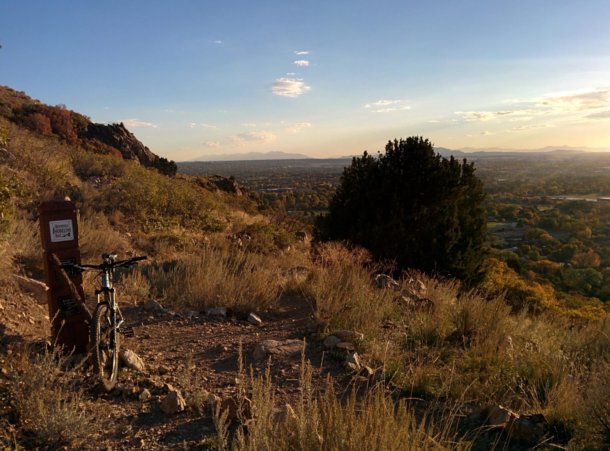 A mountain bike rests against a wooden trail sign on a dirt path surrounded by tall grass and rocks. In the background, a panoramic view of a valley and distant mountains is illuminated by the warm glow of sunset. Bonneville Shoreline Ogden North Of 12th mountain bike trail.