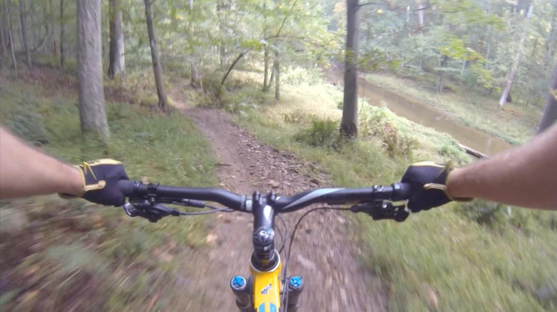 A first-person view of a mountain bike handlebar while riding on a dirt trail through a forested area, with trees and a river visible in the background. Gunpowder Falls State Park mountain bike trail.