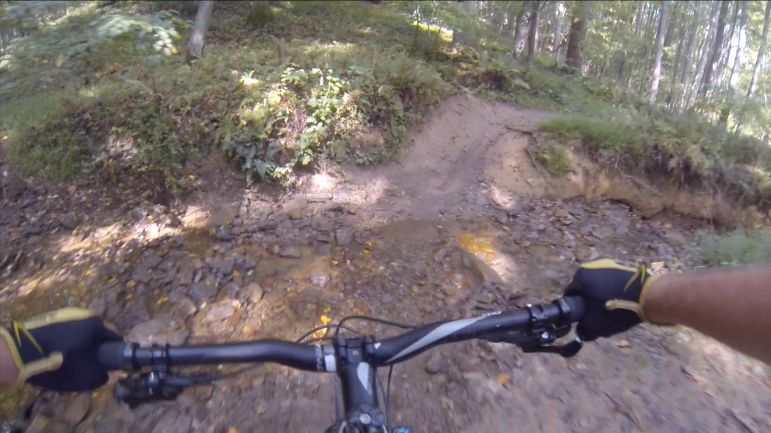 A mountain biker's perspective on a dirt trail, showcasing rocky terrain with a small creek crossing, surrounded by greenery and trees in a forested area. Gunpowder Falls State Park mountain bike trail.