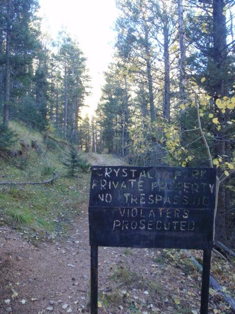 A wooden sign in a forest setting reads "Crystal Park, Private Property, No Trespassing, Violators Prosecuted." The sign is positioned along a dirt path surrounded by tall trees and underbrush, with sunlight filtering through the branches. Palmer Trail / Section 16 mountain bike trail.