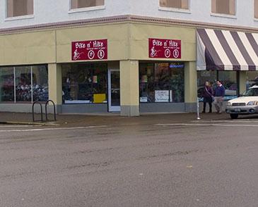 Exterior view of a bike shop called "Bike & Hike," featuring large windows displaying various bicycles and outdoor gear. The shop has a red and white sign, with two people standing on the sidewalk nearby. A parked car is visible in the foreground, along with a bike rack. The building has a striped awning above the entrance.