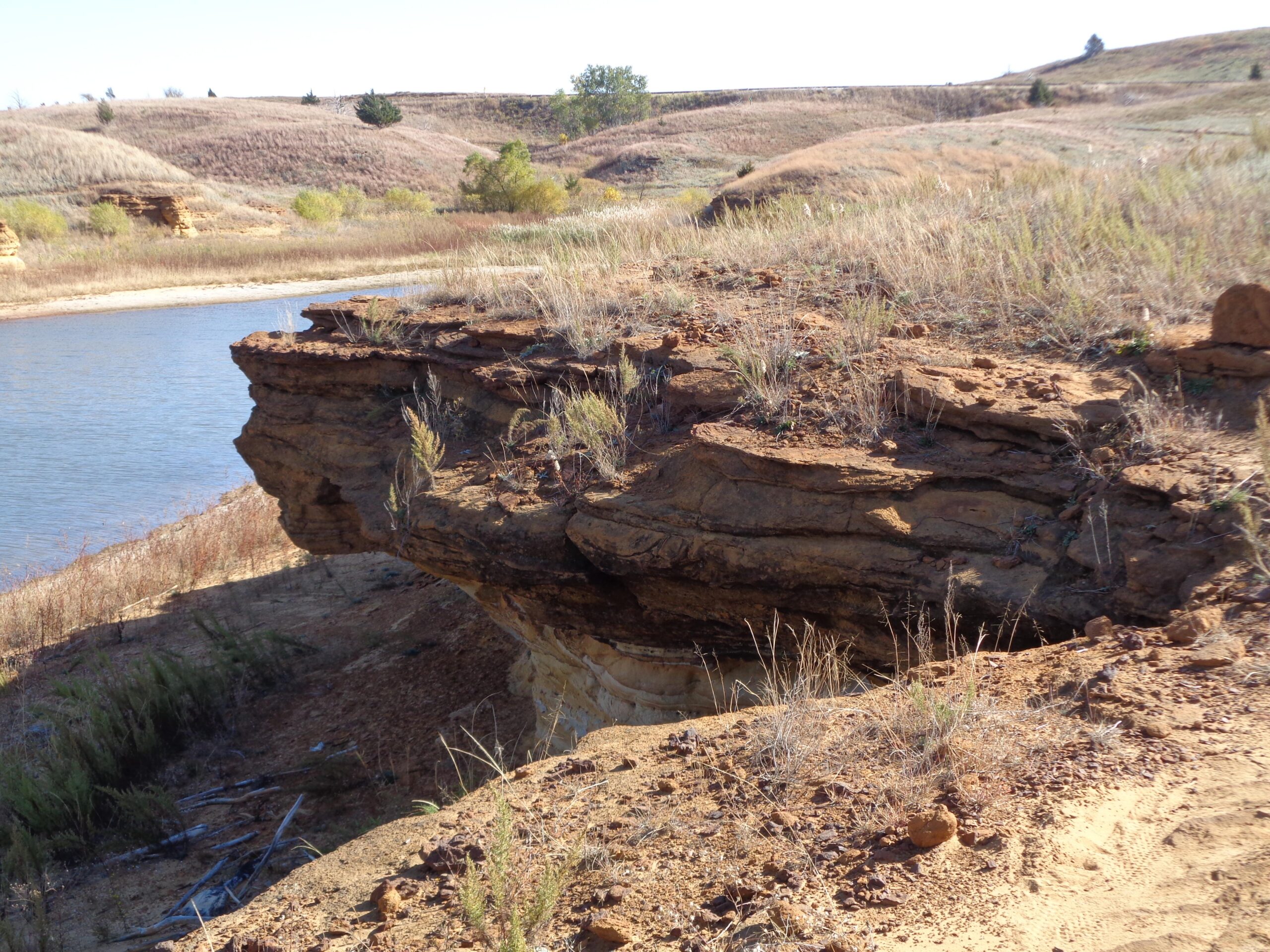 A rocky outcrop overlooking a calm body of water, surrounded by dry grass and sparse vegetation. The landscape features sandy soil and rolling hills in the background under a clear blue sky. Switchgrass mountain bike trail.