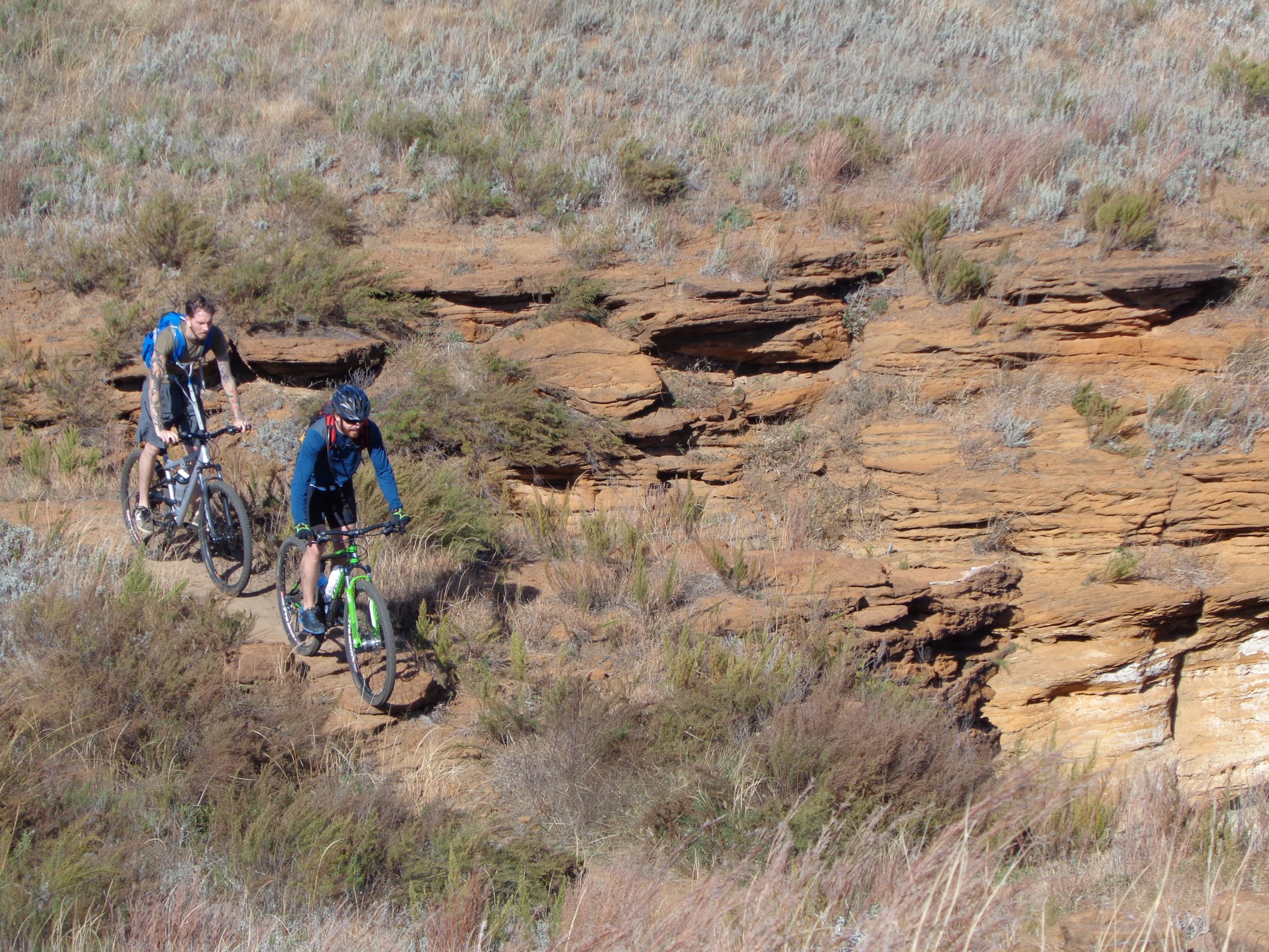 Two mountain bikers navigating a rugged trail across a rocky landscape, surrounded by tall grasses and sparse vegetation. The terrain features natural rock formations, with one rider in the foreground and the other climbing a slight incline in the background. Switchgrass mountain bike trail.