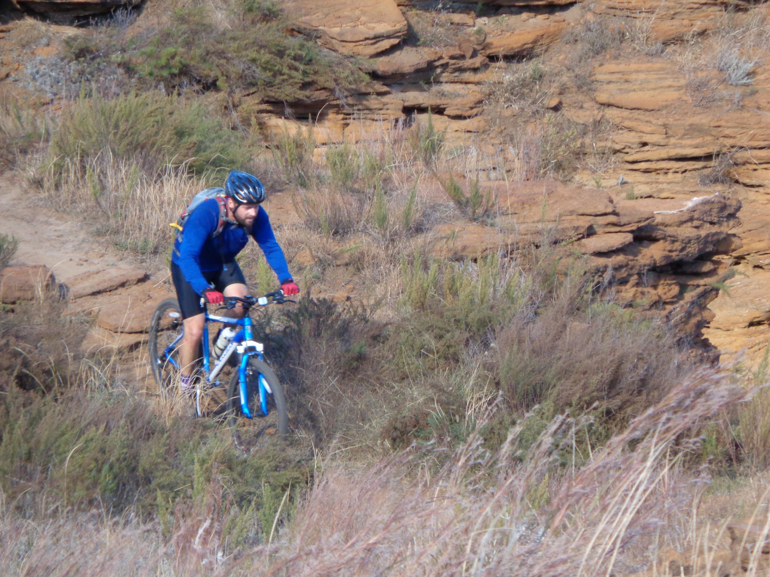 A person riding a mountain bike on a dirt trail near rocky terrain, surrounded by dry grass and shrubs. The rider is wearing a blue long-sleeve shirt, black shorts, gloves, and a helmet. Switchgrass mountain bike trail.
