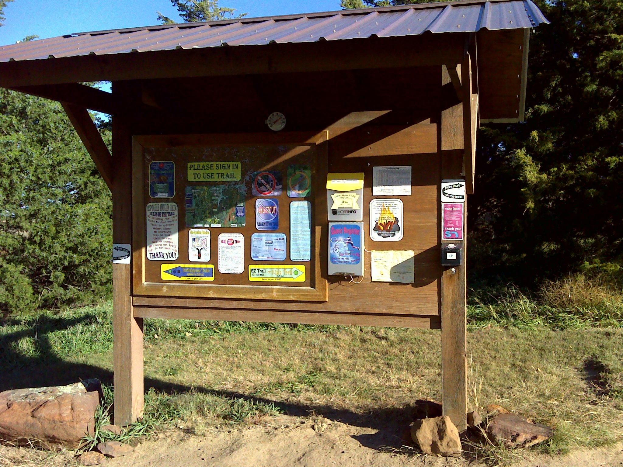 A wooden information board with a metal roof, displaying various signs and notices, including a map, rules for trail usage, and guidance for visitors. The setting features green grass and trees in the background. A sign at the top reads, "PLEASE SIGN IN TO USE TRAIL." Switchgrass mountain bike trail.