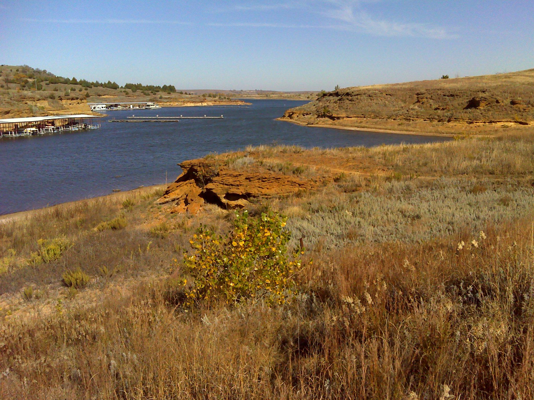 A scenic view of a calm river bordered by rocky cliffs and grassy banks. In the foreground, a small shrub with yellow leaves stands among tall grasses, while a marina with boats is visible in the background under a clear blue sky. Switchgrass mountain bike trail.