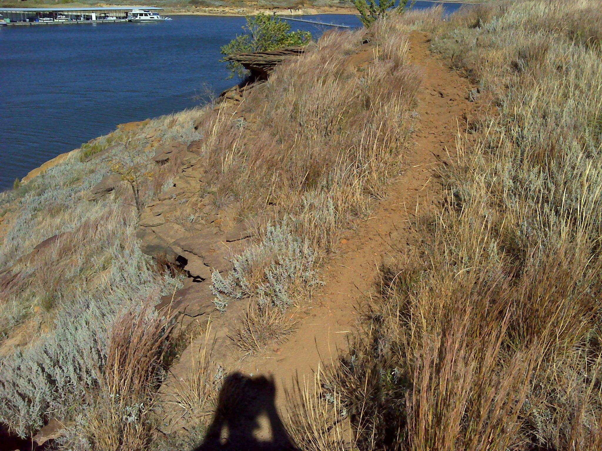 A scenic view of a dirt path leading along a hillside by a body of water, surrounded by tall grass and sparse vegetation. In the background, a docked boat is visible on the water. A shadow of a camera or person is present in the foreground. Switchgrass mountain bike trail.