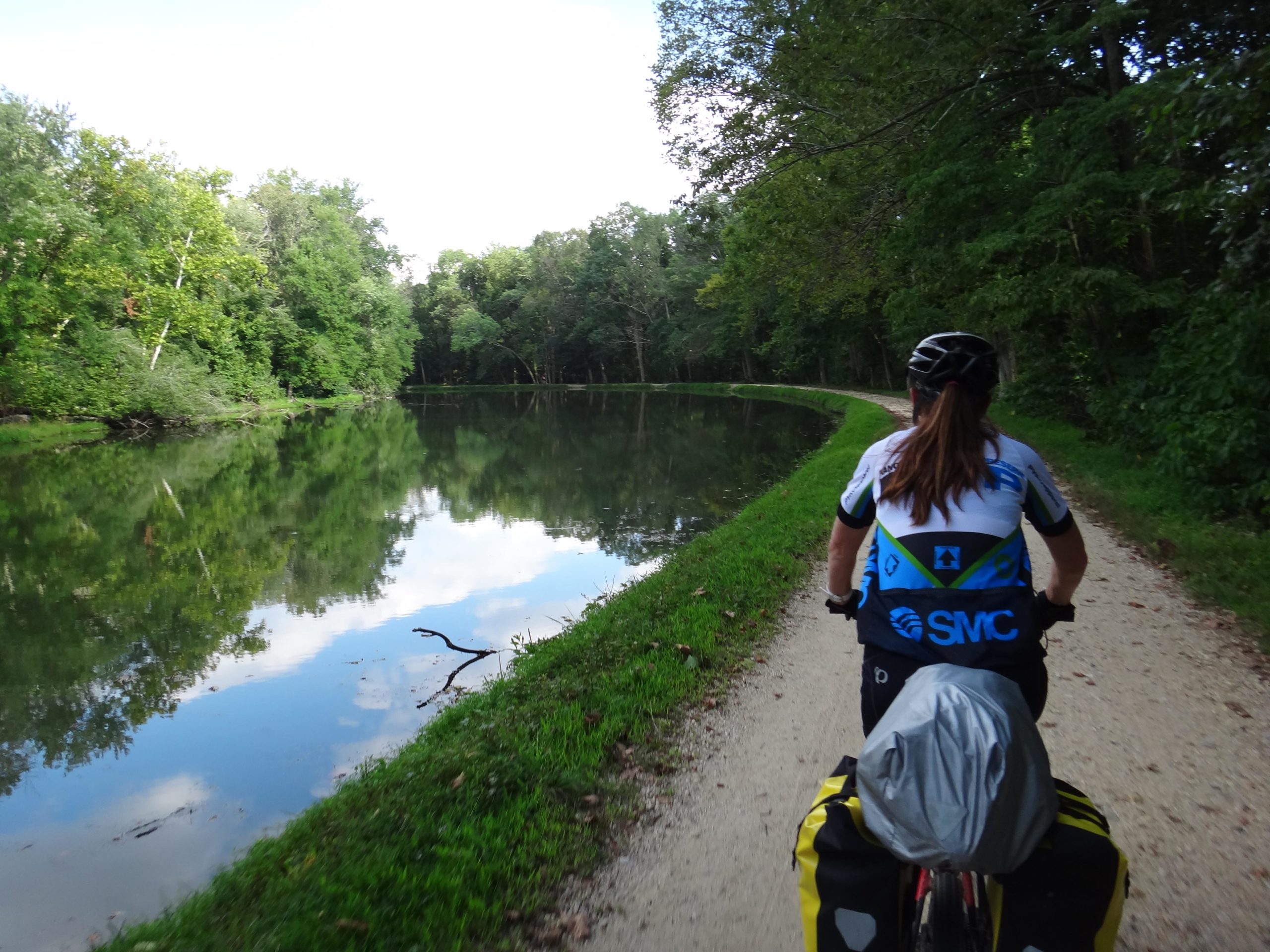 A cyclist rides along a gravel path next to a calm river, surrounded by lush greenery and trees. The reflection of the trees and sky can be seen in the water, creating a serene outdoor scene. The cyclist is wearing a blue and black jersey and has bags attached to their bike. C&O Canal mountain bike trail.