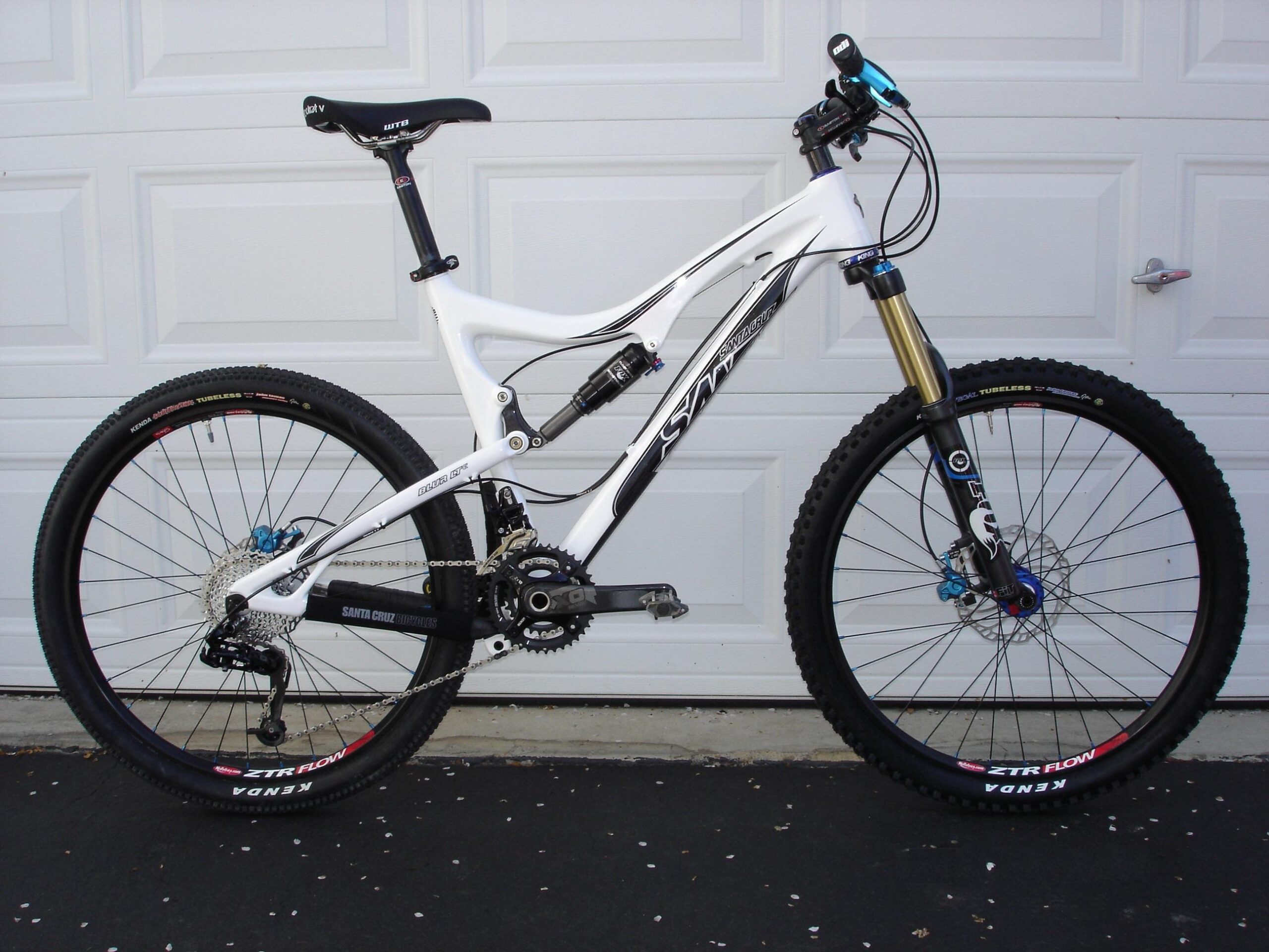 Santa Cruz Blur LT: A side view of a mountain bike featuring a white and black frame, equipped with a front suspension fork, disc brakes, and wide tires. The bike is positioned against a white garage door, highlighting its sleek design and components, including a dropper seat post and gear system.