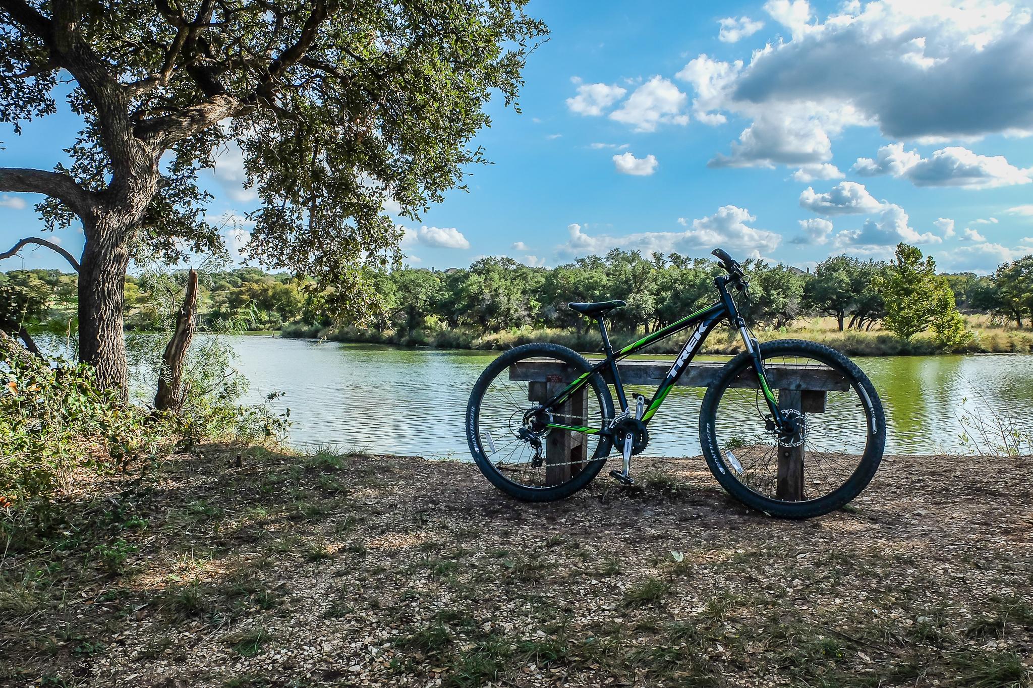 Trek Marlin 6: A mountain bike resting on a wooden post beside a calm river, surrounded by lush greenery and trees under a partly cloudy blue sky.